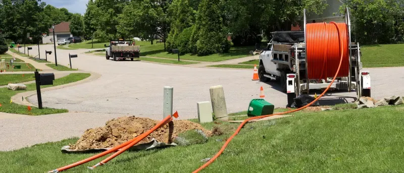 Utility workers installing underground fiber optic cables in a residential neighborhood, with orange conduit tubing laid across lawns and connected to utility boxes.